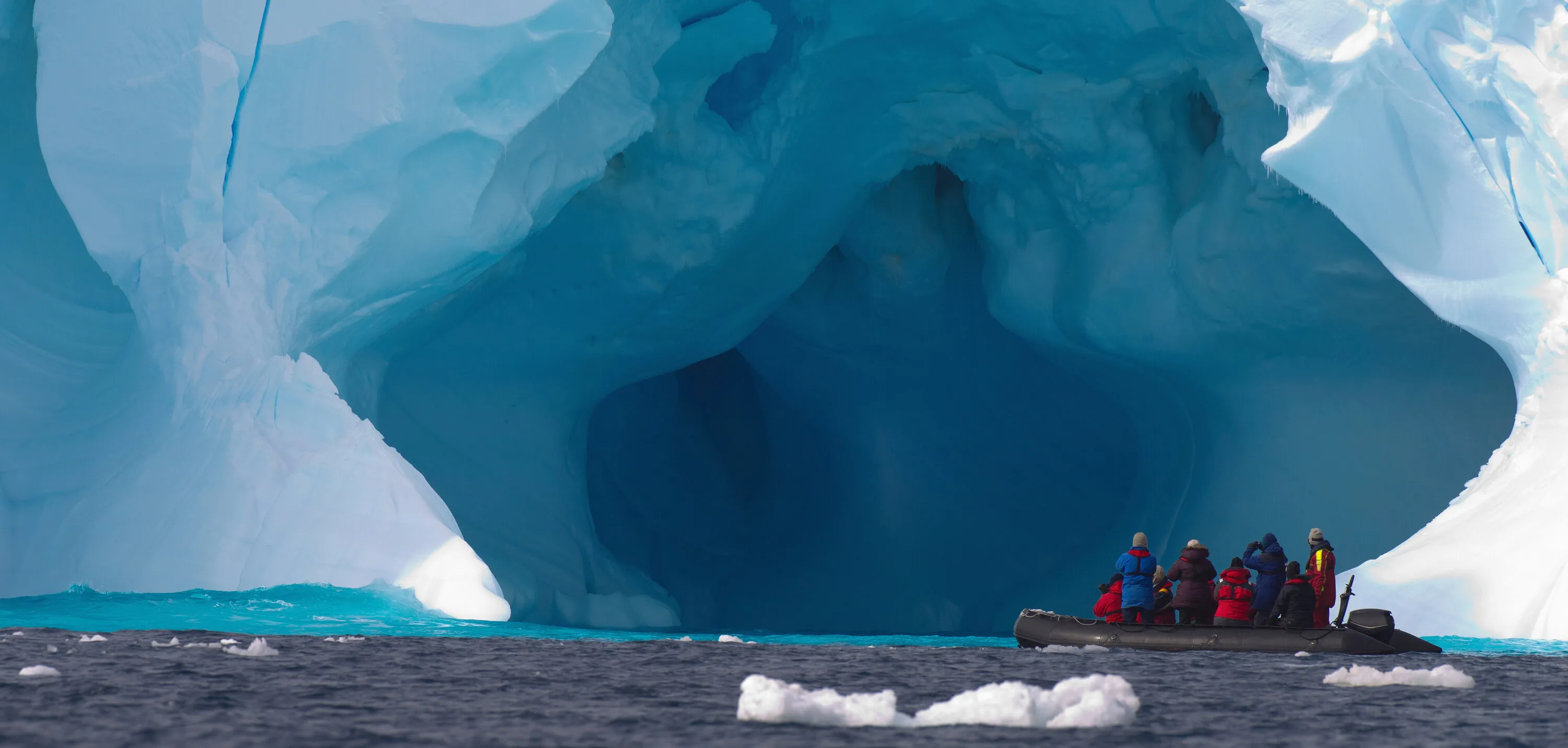 Expedition travelers in a Zodiac beside a towering blue Antarctic iceberg cave